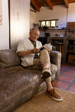 Senior Asian man sitting on brown leather sofa reading paperback book in rustic kitchen lounge