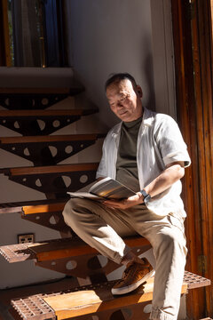 Senior Asian man sitting on wood spiral stairs reading paperback book wearing watch in sunlight