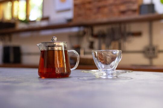 Glass teapot and clear teacup are sitting on pale tabletop, catching warm left light