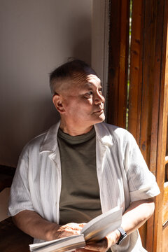 Senior Asian man sitting by wooden window, wearing olive shirt, holding open book, watch visible
