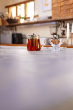 Glass teapot is steeping tea on pale countertop with empty teacup and sunlit window, copy space