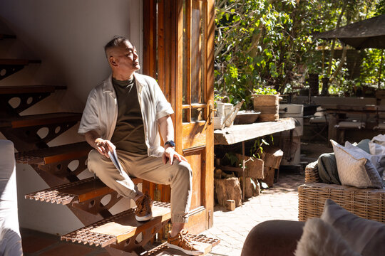Senior Asian man sitting on open-tread metal stairs by open wooden door, reading paperback notebook