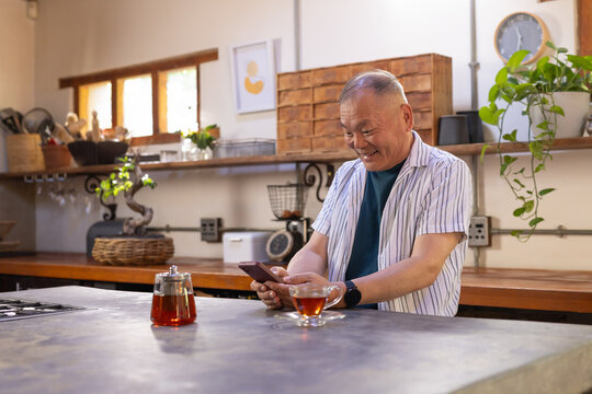 Senior Asian man standing at home kitchen counter, holding phone and glass teacup, in striped shirt