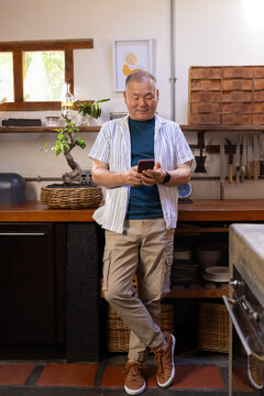 Senior Asian man leaning against wood countertop in kitchen, checking smartphone with bonsai basket