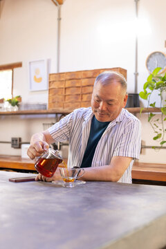 Senior Asian man pouring tea from glass teapot into cup at counter with smartphone, striped shirt