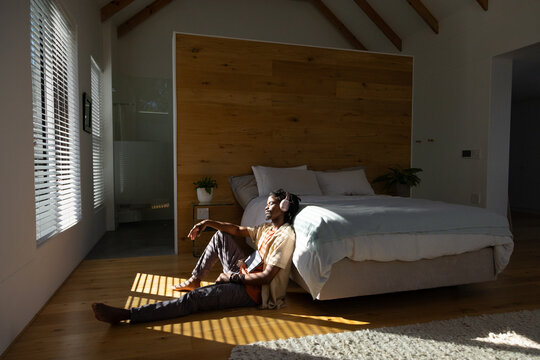 African American man sitting on floor in bedroom by bed, wearing headphones and holding phone