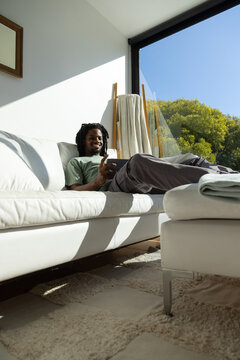 African American man reclining on white leather sofa by floor-to-ceiling window, holding tablet
