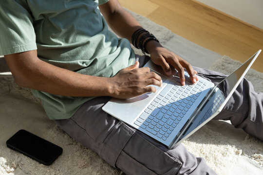 African American man wearing green tee working on laptop on beige rug with smartphone, bracelets