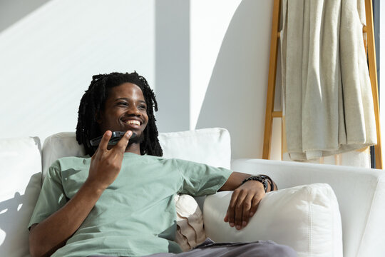 African American man reclining on sofa wearing green tee holding phone near mouth, ladder behind