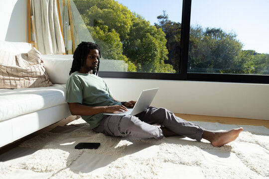 African American man sitting in living room on rug using silver laptop with black smartphone nearby