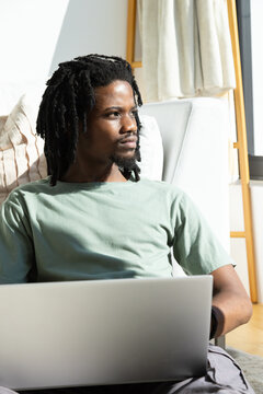 African American man in 20s sitting in green T-shirt on armchair by window, working on laptop
