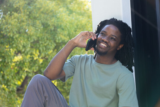 Adult African man sitting at white-column on step, talking on smartphone in green tee, copy space