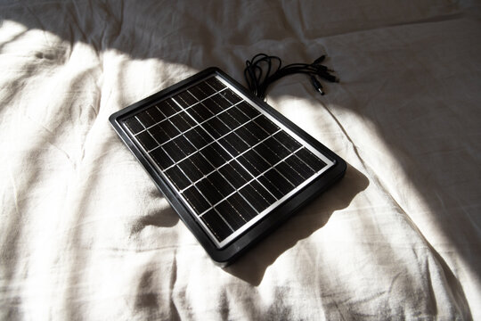 Portable solar panel is lying diagonally on cream bed sheet with coiled cables and sharp shadow