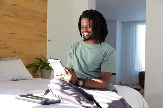 Adult African American man sitting cross-legged on bed in bright bedroom holding tablet and folio
