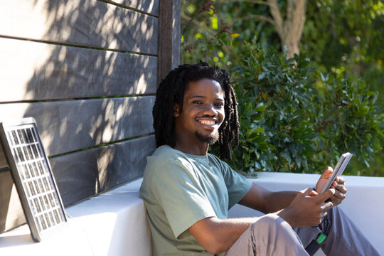 Adult African man sitting on bench in courtyard wearing green tee, holding tablet, solar panel