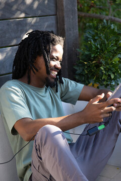 African American man sitting on patio in light green tee holding tablet with inline adapter