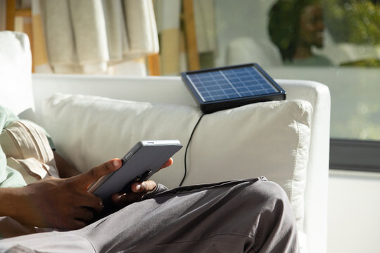 Gray tablet is charging from portable solar panel while resting on white sofa near sunlit window