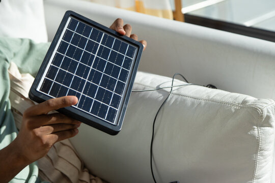 Adult African American man wearing green shirt holding mini solar panel on leather sofa with cable