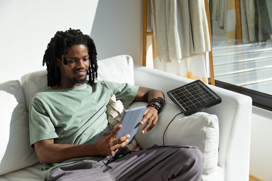 African man sitting on white couch near large window, holding tablet and charging with solar panel