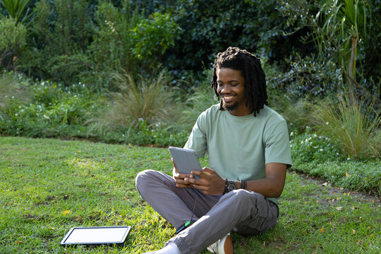 Man sitting cross-legged on garden lawn wearing green tee reading tablet with second tablet left