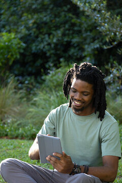 African man sitting on park grass tapping tablet with cord wearing light green tee watch bracelets