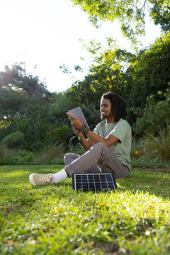 Man sitting cross-legged on grass in wooded park charging tablet with solar panel, copy space