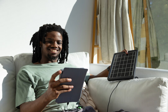 African American man wearing green T-shirt sitting on couch at home holding tablet and solar panel
