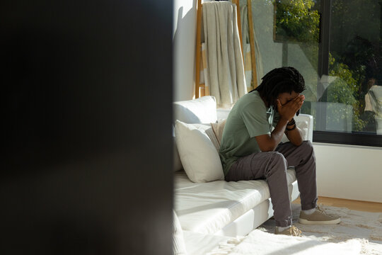 African adult male sitting on sectional sofa near window, covering face, ladder and pillows visible