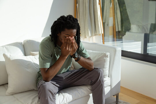 Adult African man sitting on white sofa covering face with hands by glass window, wearing bracelets