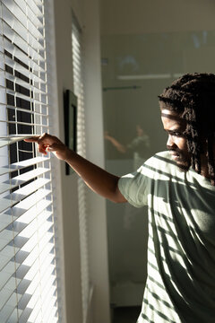 Man standing in bathroom lifting blind slat, peeking through blinds at sunlight stripes, copy space