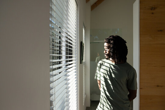 Adult African man standing, gazing through window blinds toward glass panel reflection, copy space