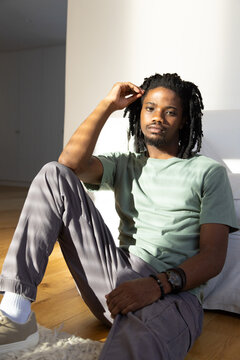 Man sitting on hardwood floor near sofa in bright apartment touching hair, wearing bracelets