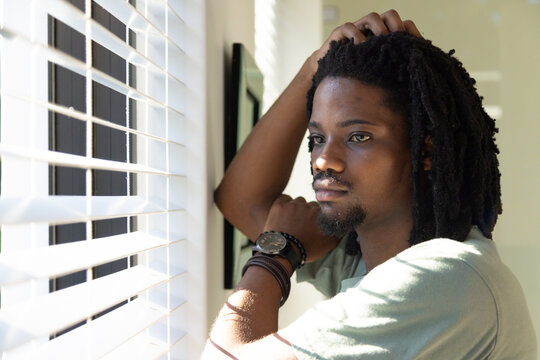 African American man leaning by window with white blinds, touching hair, showing watch, copy space