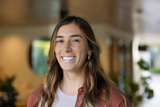 Woman standing and smiling in modern office lobby, wearing rust button-up shirt and earrings