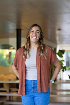 Woman standing, smiling at camera at home wearing rust shirt, jeans, wristwatch, earrings