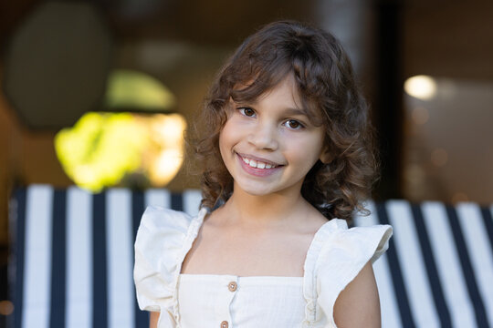 African American girl smiling, sitting on striped bench at porch, wearing white buttoned dress