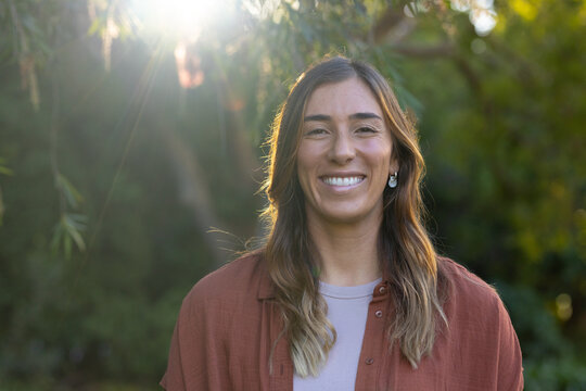 Adult woman standing outdoors in garden, smiling, wearing rust-colored overshirt and drop earrings