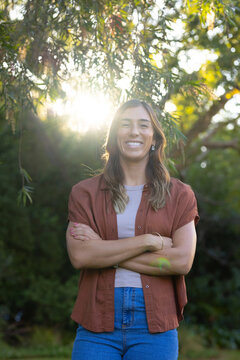 Adult woman standing smiling in park with trees, sunlight causing lens flare, in rust-brown shirt