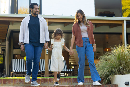 Diverse family walking down brick steps at front of modern house, parents holding child's hands