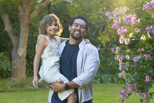 African American father standing on lawn holding smiling daughter in light dress, watch, pink shrub