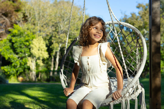 African American preteen girl sitting on macrame chair in garden holding rope in cream romper