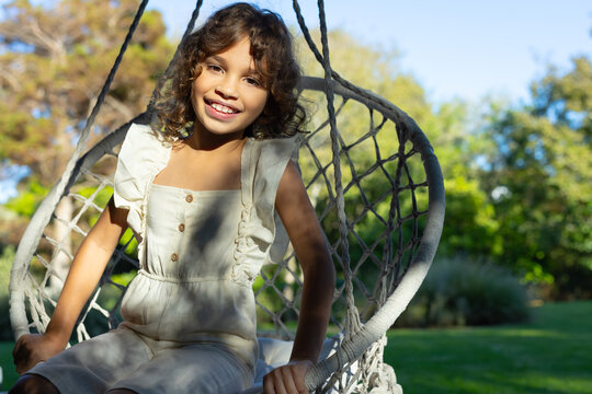 Female child sitting rope swing in garden holding edges, linen romper with wooden buttons, smiling