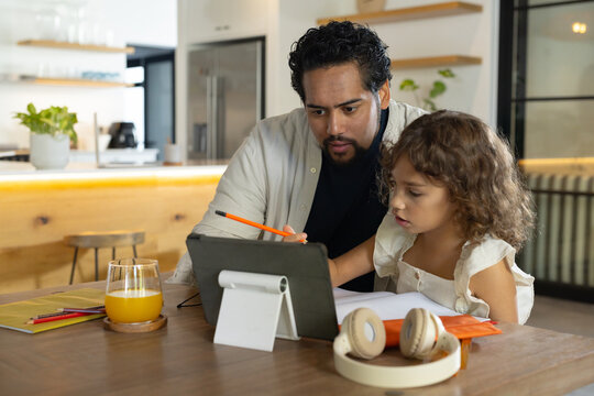 African American father and daughter sitting at wooden kitchen table using tablet and orange pencil