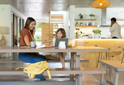 Diverse family sitting at table in open-plan kitchen, woman holding mug, child using tablet nearby