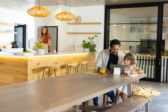 Diverse family sitting at open-plan kitchen table using tablet, child eating bowl, copy space