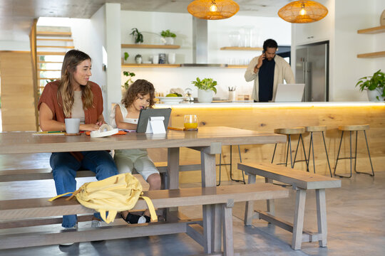 Diverse family sitting at kitchen table using tablet, yellow backpack, father calling, copy space