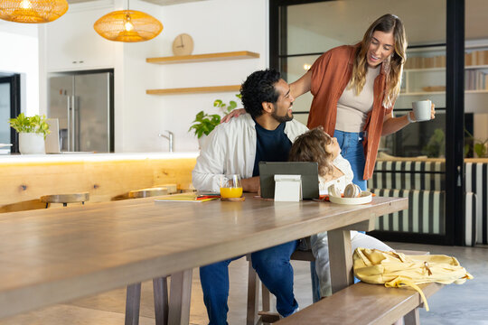 Family with child sitting at long wooden table at home, parents leaning, holding mug, tablet nearby