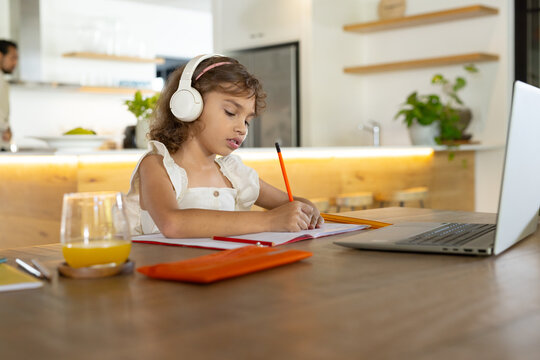 African American child with adult writing at table in kitchen, using laptop and headphones