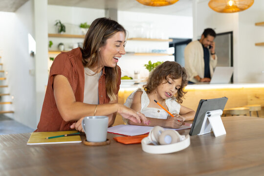 Diverse family woman leaning toward child, helping write with pencil and tablet at kitchen table