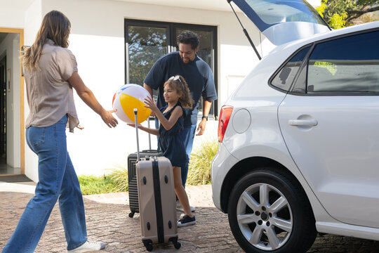 Diverse parents with child packing suitcases loading white hatchback on driveway with beach ball